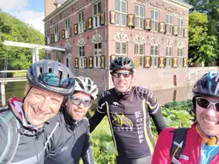 Vibrant group of cyclists smiling outdoors near a historic brick building, enjoying a sunny day for outdoor activities and team-building events.